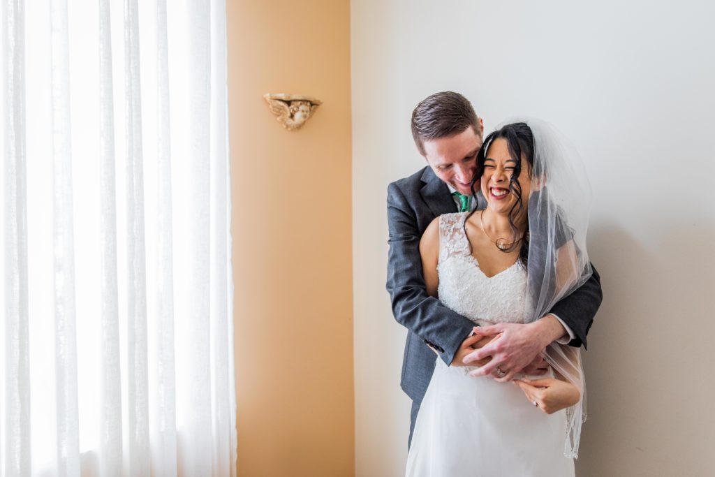 Groom holds bride closely by a window