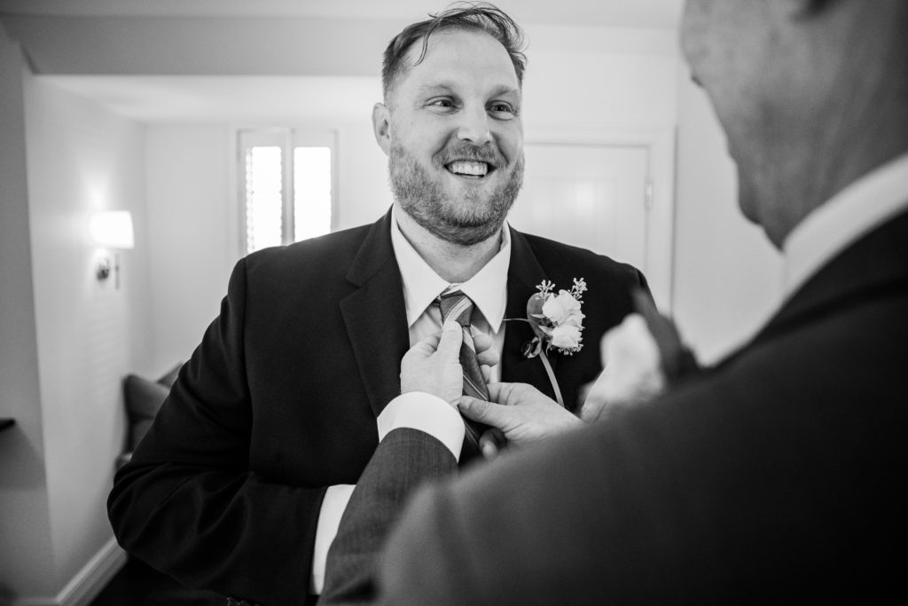 Father of the groom adjusting the groom's tie while he smiles