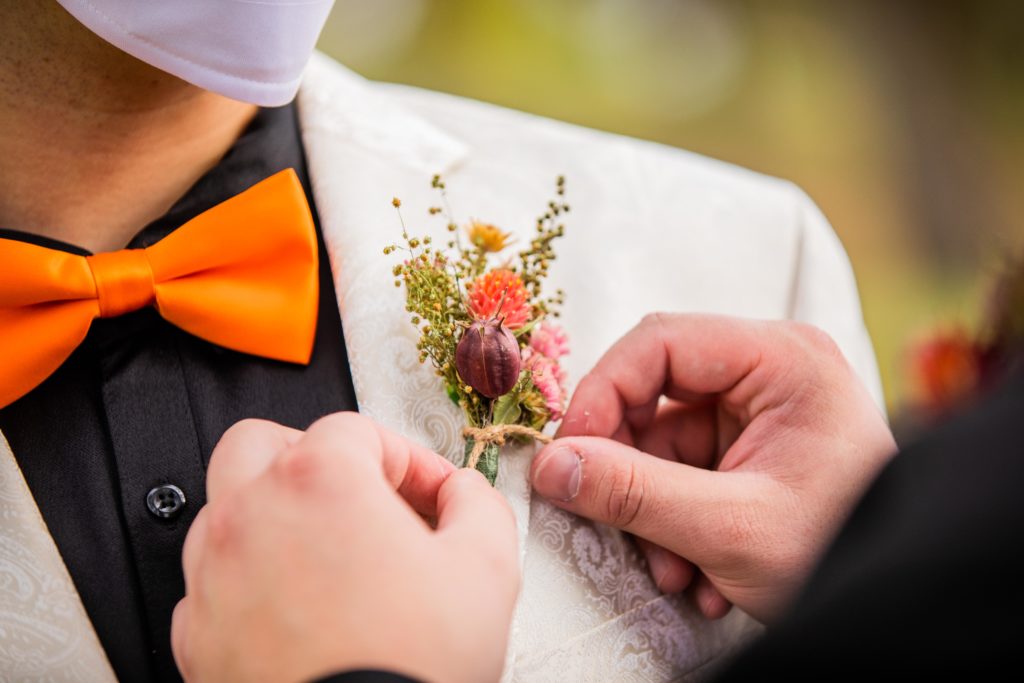 Man puts boutonniere on his husband while wearing a mask 