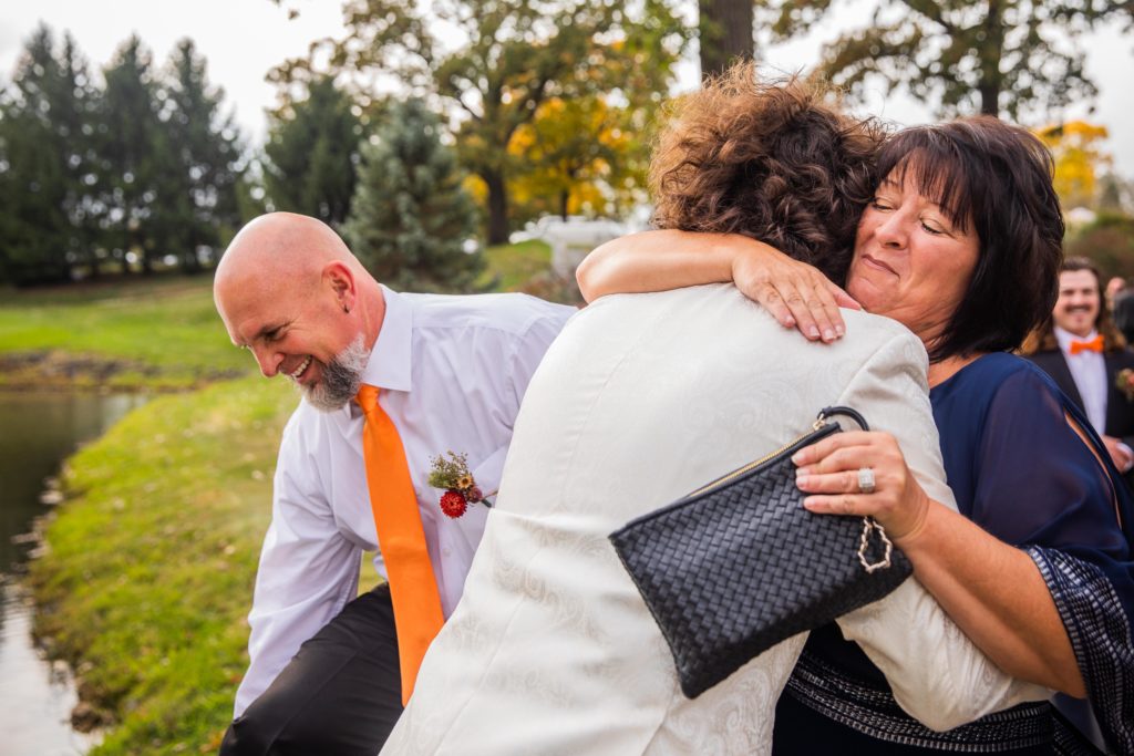 Mother of the groom hugs his husband while father smiles in the background