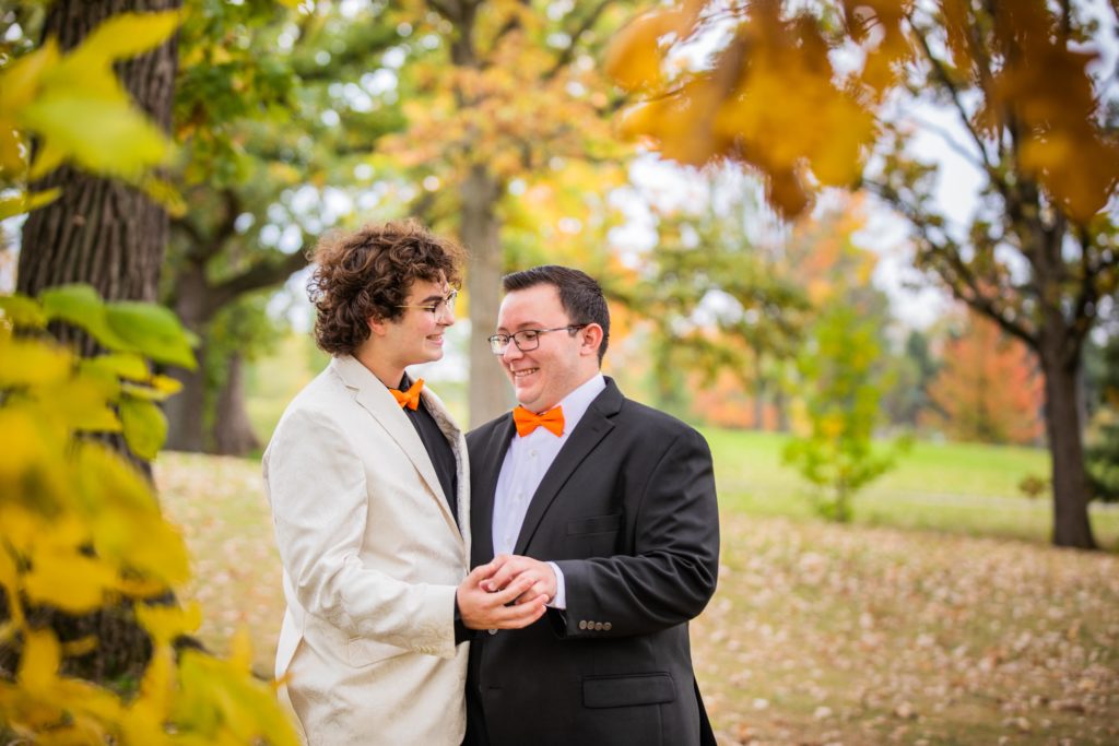 Grooms smiling and holding hands behind yellow leaves at the Silver Lake Country Club