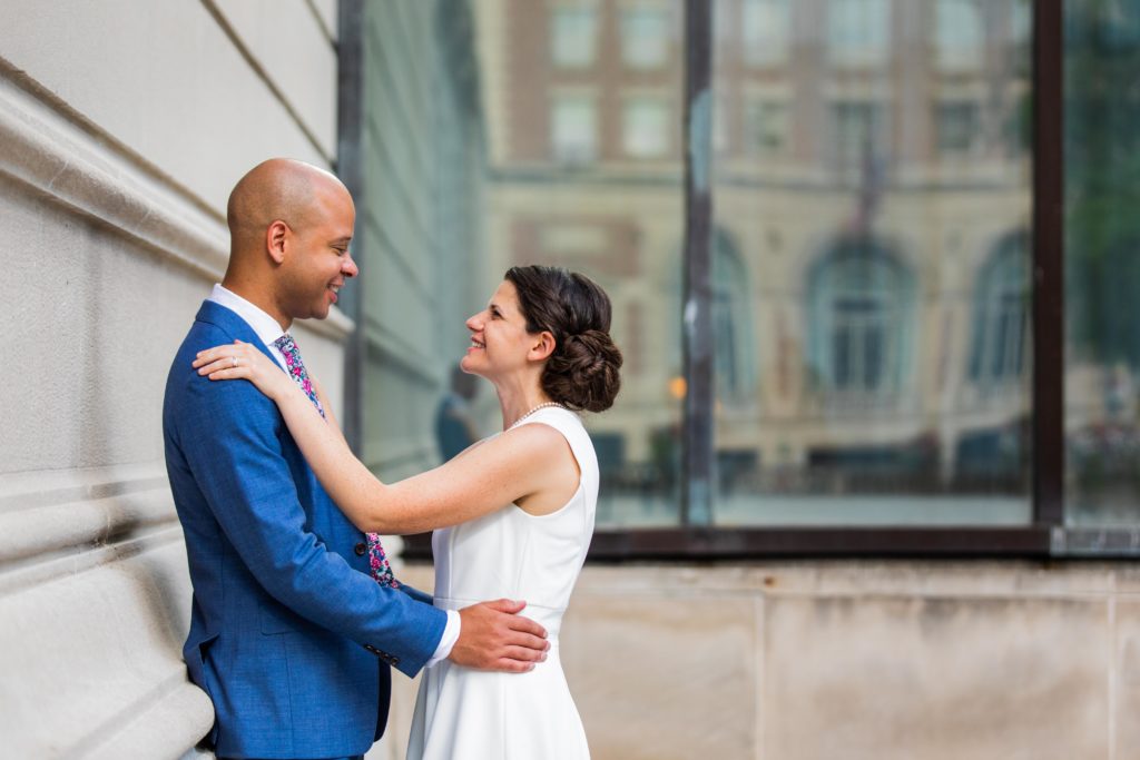 Bride and groom smile at each other while the groom leans against a wall of the Art Institute