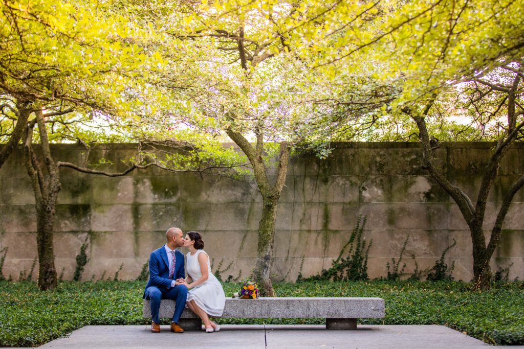Bride and groom kiss on a bench underneath trees
