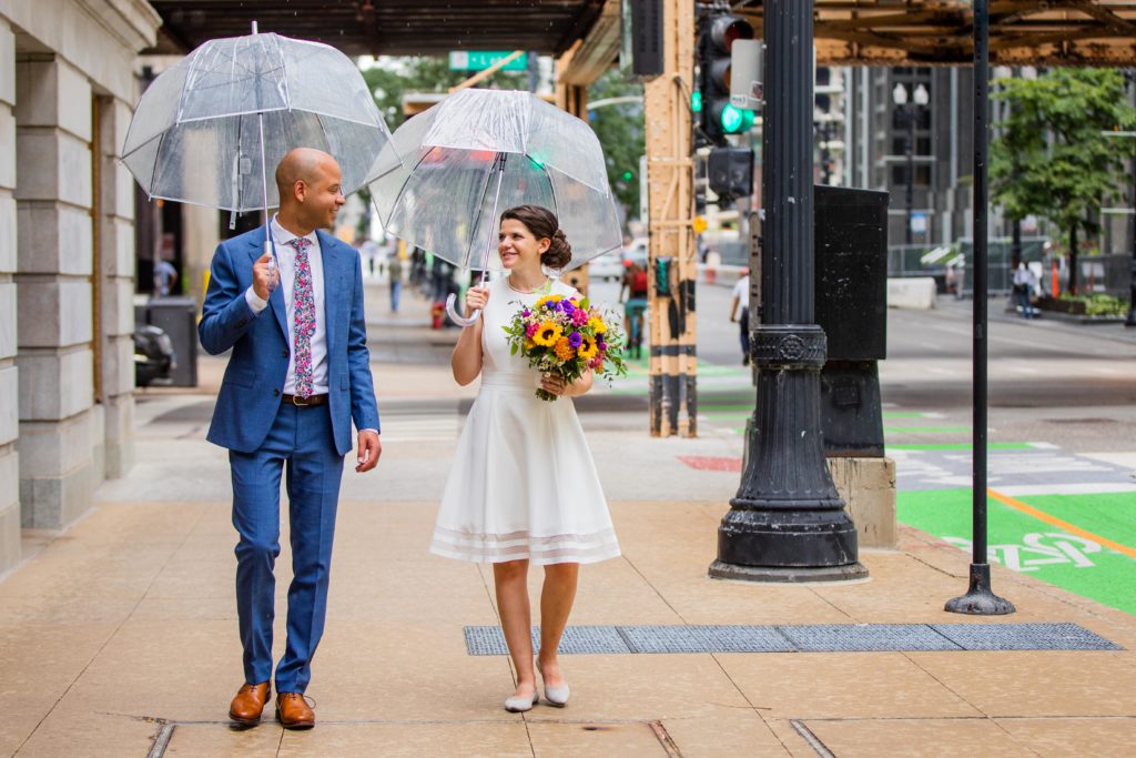 Bride and groom walk on the sidewalk in downtown Chicago while holding umbrellas