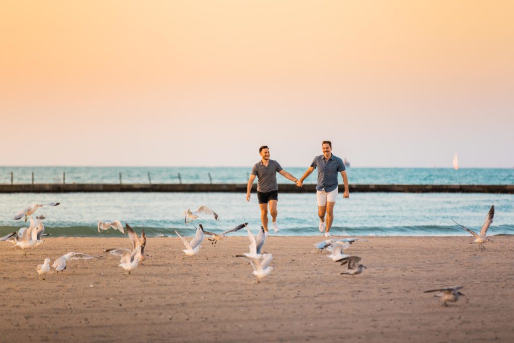 Men running hand in hand into the sunset together on the beach with seagulls in the background at North Avenue Beach