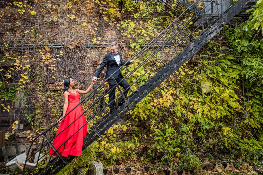 Groom leading the bride in a red dress up a fire escape leaning on a wall covered in vines