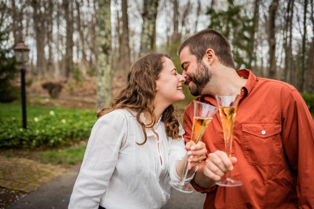 Couple clink champagne flutes while leaning in for a kiss during their engagement session in Bedford