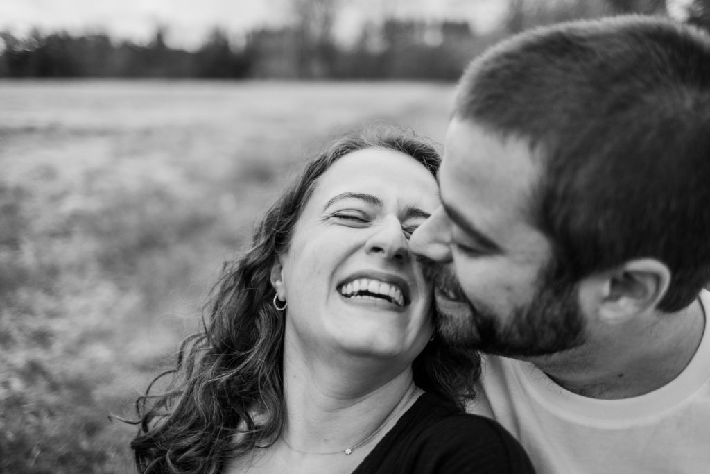 Woman laughs as man leans in to kiss her during their engagement session in Bedford