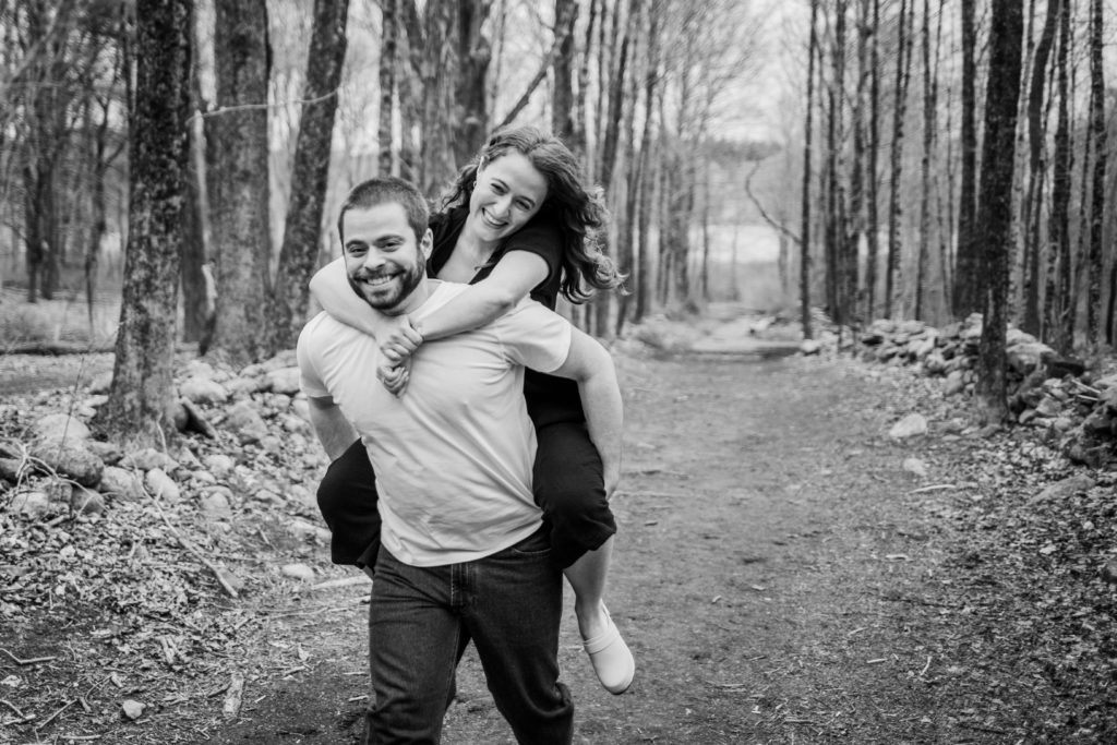 Couple laugh as the man runs through the woods giving a woman a piggyback ride during their engagement session in Bedford