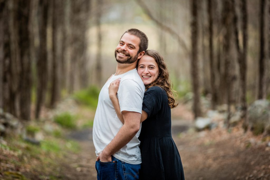 Couple smile while woman embraces the man from behind during their engagement session in Bedford