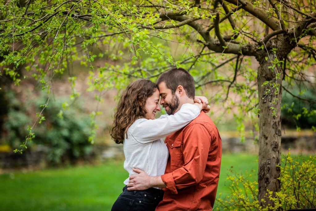 Man holds woman's hips while she wraps her hands around his neck while they touch foreheads during their engagement session in Bedford