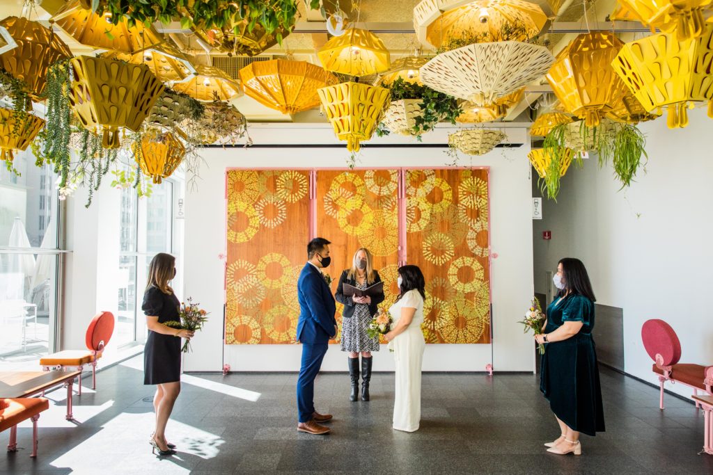 Bride and groom face each other with witnesses and officiant in a back room of a museum