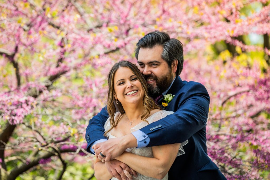 Man holds woman from behind as she smiles in front of a pink tree