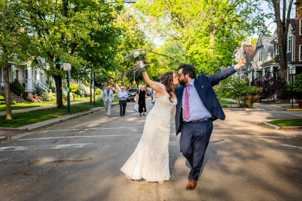 Bride and groom kiss while holding up their fists in the middle of the street on the way to their Reclaimed Bar and Restaurant Wedding reception