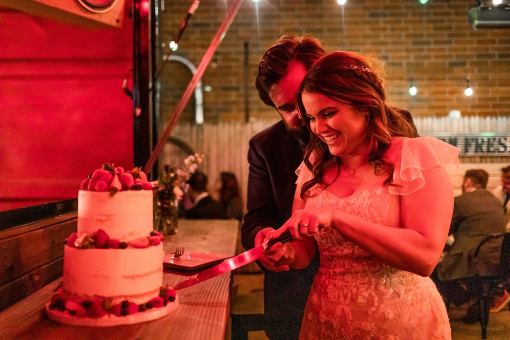Couple smile while cutting their cake from Alliance Bakery during their Reclaimed Bar and Restaurant Wedding reception