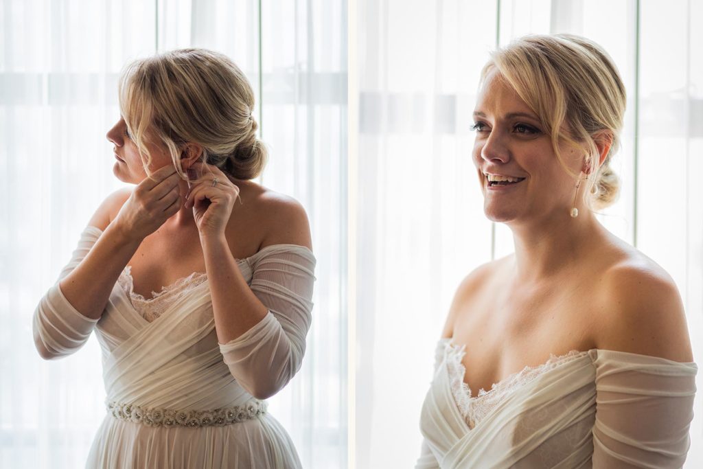 Bride smiling and putting on her earrings in front of a window at the Drake