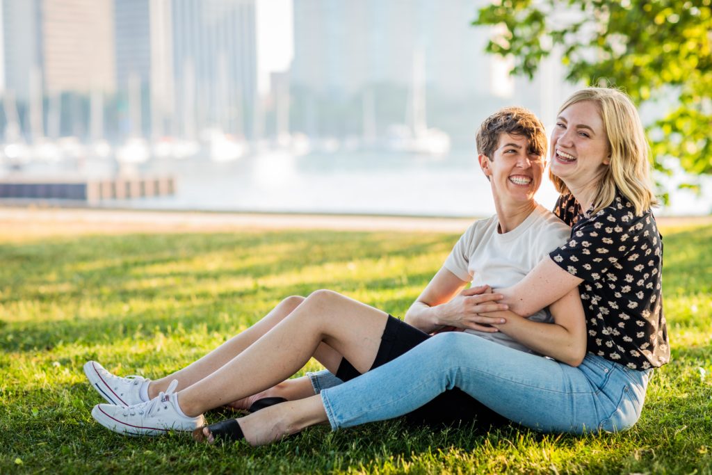 Woman embracing another woman from behind while sitting on the grass during their engagement session at belmont harbor