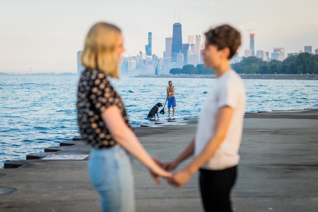 Women holding hands while a paddle boarder moved behind them with a dog on the board during their engagement session at belmont harbor