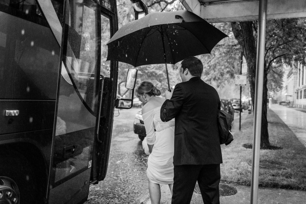 Bride gets into a bus in the pouring rain as someone holds an umbrella