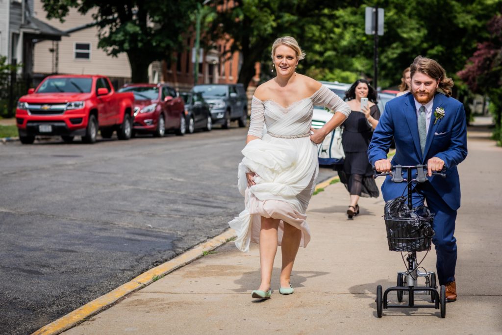 Bride walks next to the groom while he rolls down the sidewalk on a scooter before their Big Star Reception