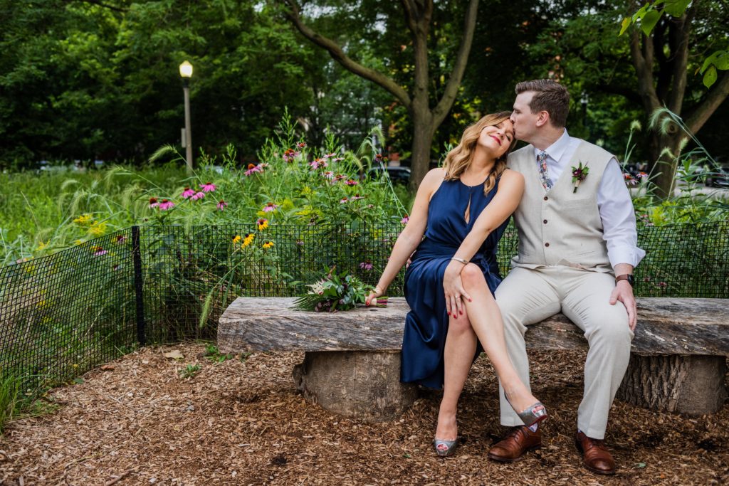 Groom kissing bride on the forehead while they sit on a bench