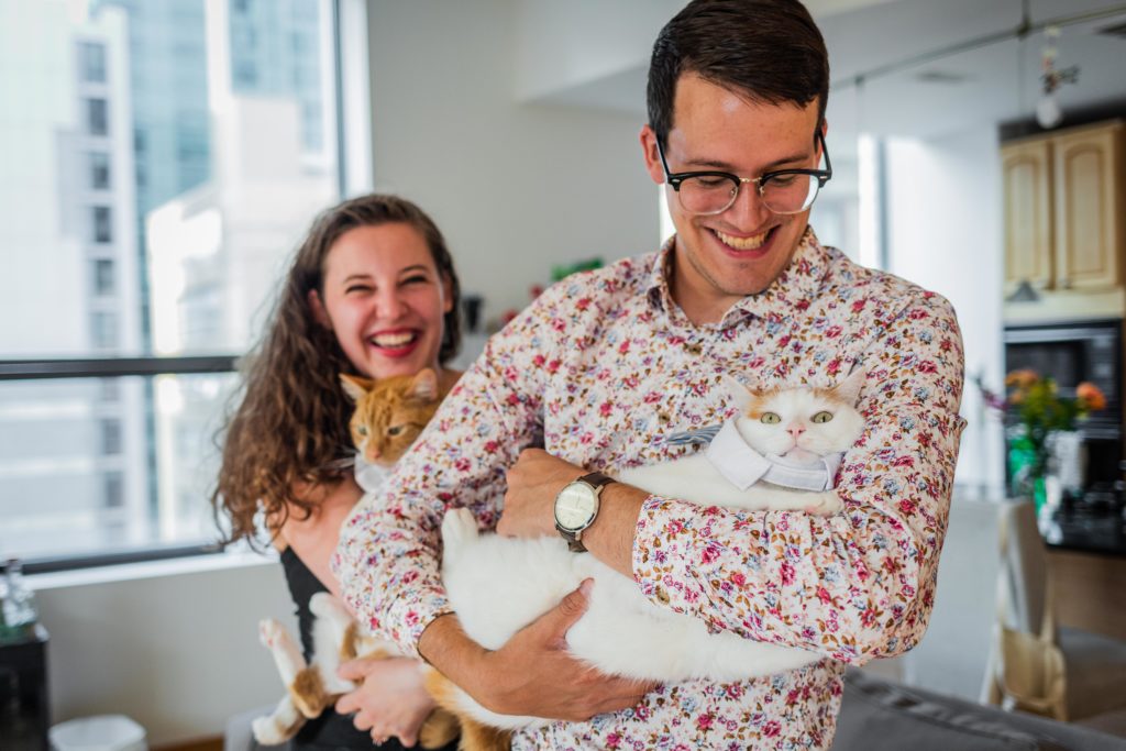 Couple laughing and holding cats while the cats try to squirm out of wearing clothes