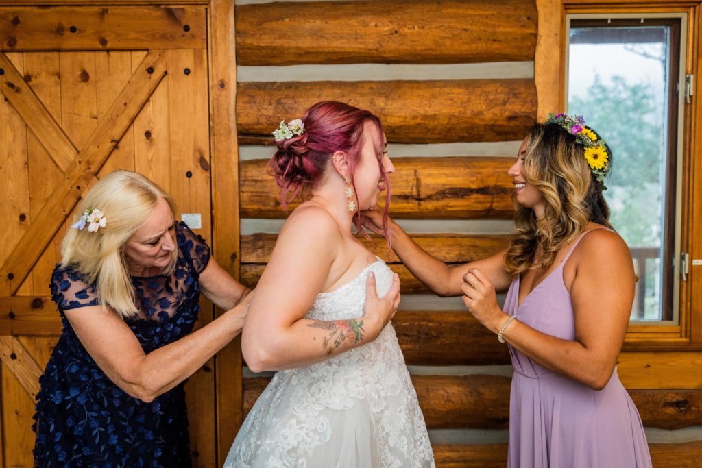Mother of the bride helping the bride put on her dress while the maid of honor helps
