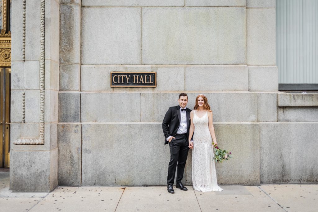 Couple standing and smiling in front of the City Hall sign in Chicago