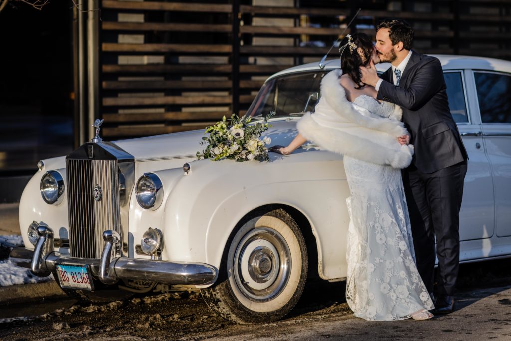 Groom kisses bride while they kiss and lean on a vintage car before their wedding at Ovation Chicago